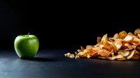 Fresh green apple contrasts with pile of crispy dried apple chips on dark background, highlighting healthy versus processed snack options.