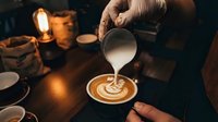 Barista pouring steamed milk creating latte art in coffee cup at cozy cafe with warm lighting and rustic atmosphere for hospitality and beverage service.