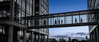 Modern office buildings connected by glass walkway bridge with business people silhouettes walking across, urban corporate architecture against blue sky.