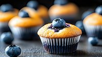 Fresh blueberry muffin with ripe berries on dark wooden background, selective focus on single muffin garnished with whole blueberry on top.