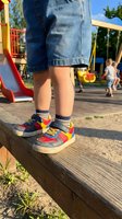 Child wearing denim shorts and colorful sneakers sitting on wooden beam at playground with other children playing in background on sunny day.