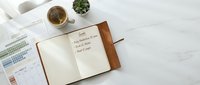 Open leather journal with handwritten goals on white marble desk alongside coffee cup, calendar and succulent plant for productivity planning.