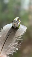 Miniature wooden house with moss balanced on feather tip against blurred natural background symbolizing lightness and fragility of home dreams.