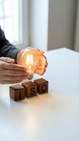 Hands holding a glowing Edison bulb beside wooden blocks spelling ISO on a white desk, symbolizing quality standards and innovation.