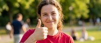 Happy young Caucasian woman with curly hair giving thumbs up gesture in sunny park wearing red shirt, positive attitude and success concept for lifestyle content.