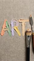 Word "ART" spelled with colorful paint strokes in pastel shades on neutral linen background, accompanied by professional art brushes and palette knife for creative projects.