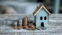 Small wooden house model with stacks of coins on rustic surface representing real estate investment, mortgage savings, property finance and home buying concept.