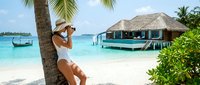 Woman in white swimwear and hat relaxing on tropical beach with overwater bungalows, turquoise lagoon and palm trees for travel and vacation concepts.
