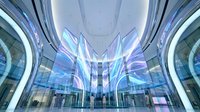 Young woman with light skin tone, professional appearance, wearing business casual attire, standing in modern glass and steel lobby with symmetrical architecture, blue and purple lighting, contemporary corporate environment.