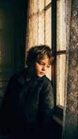 Young Caucasian man with tousled hair sitting pensively by vintage window with warm golden sunlight streaming through glass panes in moody atmospheric interior.