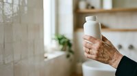 Senior woman holding white cosmetic bottle in modern bathroom with natural light and wooden shelving.