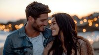 Young romantic couple sharing intimate moment at sunset with bokeh city lights in background, expressing genuine happiness and connection in casual attire.