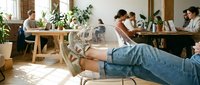 Young woman with tattoos relaxing in modern coworking space while diverse team collaborates on laptops at wooden tables in bright office environment.