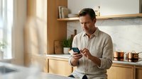 Middle-aged Caucasian man in casual beige shirt using smartphone in modern kitchen with wooden cabinets and marble backsplash, natural lighting from window, home technology and communication concept.