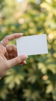 Woman's hand holding a blank white business card against a blurred green bokeh nature background, mockup for branding and design.