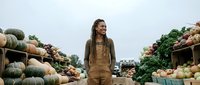 Young African American woman in overalls standing among fresh produce at farmers market with pumpkins and vegetables for healthy lifestyle content.