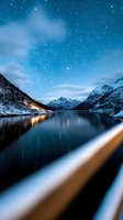Starry night sky over snow-covered mountains and frozen alpine lake with cabin lights reflecting on water. Winter landscape for travel, nature, and astronomy projects.