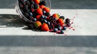 Fresh mixed berries spilling from wicker basket on concrete surface. Strawberries, blueberries, blackberries scattered creating colorful summer harvest scene.