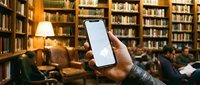 Hand holding smartphone with blank white screen in cozy library setting with wooden bookshelves and warm lighting for mockup and app presentation purposes.
