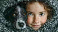 Young girl with blue eyes and mixed breed dog cuddling together in cozy gray knit blanket showing friendship bond between child and pet companion.