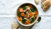 Hearty vegetable soup with carrots, beans, and leafy greens served in ceramic bowl with crusty bread on white marble surface for healthy comfort food recipes.