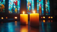 Two glowing candles against blurred stained glass windows in church, creating peaceful spiritual atmosphere with warm candlelight and colorful bokeh reflections.