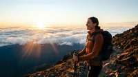 Woman hiker with backpack standing on mountain peak above clouds at sunrise, enjoying scenic golden hour landscape view during outdoor adventure trek.