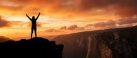 Silhouette of person with raised arms celebrating success on mountain cliff edge during golden sunset with dramatic orange sky and canyon landscape below.