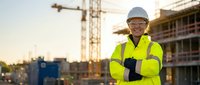 Confident female construction worker in safety gear smiling at building site with cranes and scaffolding during golden hour sunset for workplace safety campaigns.