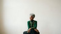 Senior woman with short gray hair wearing green blazer and gold necklace, sitting against neutral background, professional portrait for business, corporate, and lifestyle design projects.