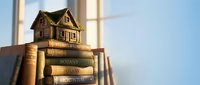 Miniature wooden house model sitting on stack of old books near bright window creating warm educational atmosphere for real estate learning concepts.
