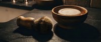 Traditional wet shaving setup with wooden bowl containing cream and natural bristle brush on dark stone surface for grooming and self-care routines.