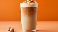 Layered iced latte with whipped cream and cinnamon in tall glass on orange background, garnished with chocolate pieces. Fresh cold coffee drink.