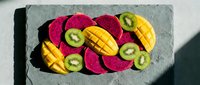 Sliced tropical fruits arranged on a dark slate board, featuring dragon fruit, mango, and kiwi in vibrant colors, top view, healthy eating concept.