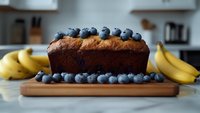 Fresh baked banana bread topped with ripe blueberries on wooden cutting board, surrounded by fresh fruit in modern kitchen setting. Homemade pastry.