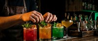 Professional bartender preparing colorful craft cocktails with garnishes at upscale bar counter with bottles and bar tools in atmospheric lighting.