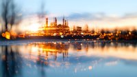 Industrial refinery plant illuminated at dusk with golden lights reflecting on water surface, creating dramatic atmospheric scene with bokeh effect and blurred foreground.