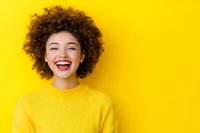 Joyful young African American woman with natural curly hair laughing with closed eyes against vibrant yellow background, wearing matching yellow sweater.