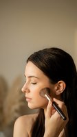 Young brunette woman applying makeup with powder brush on cheek in natural lighting for beauty routine and cosmetic application.