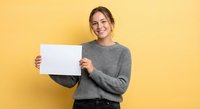 Young Caucasian woman in gray sweater holding blank white paper sheet against yellow background, smiling at camera for advertising mockup presentations.