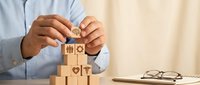 Professional businessman in light blue shirt building wooden blocks with business icons including gears, people, and hearts representing teamwork and strategy.