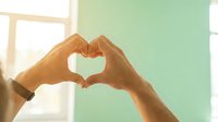Hands forming a heart shape against a bright window, symbolizing love, care, and connection in a warm, sunlit indoor setting.