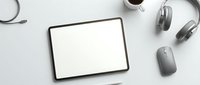 Digital tablet with blank white screen surrounded by wireless headphones, computer mouse and coffee cup on clean white desk for modern workspace mockup.