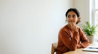 Young South Asian woman in rust-colored sweater sitting at desk with laptop, hand near mouth in thoughtful pose, contemplative expression, bright natural light from window, potted plant background.