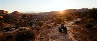Young male hiker crouching on rocky desert trail at golden hour sunset, surrounded by arid landscape with mountains and sparse vegetation for travel and adventure content.