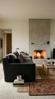 Modern living room with black curved sofa, concrete fireplace with active flames, wooden coffee table, woven rugs, and minimalist decor for interior design inspiration.
