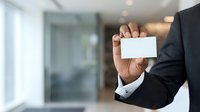 Businessman in dark suit holding blank white business card in modern office environment with blurred background, suitable for corporate branding and professional networking materials.