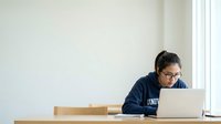 Young South Asian woman wearing glasses and dark navy hoodie, focused on laptop work at wooden desk in bright, minimalist office space with white walls and natural light from window.