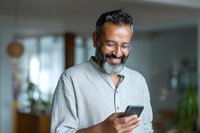 Happy mature Middle Eastern businessman with beard and glasses using smartphone in modern office, smiling while texting or browsing social media apps.