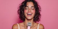 Young woman with curly hair singing into microphone against pink background, expressing joy and passion for music performance.